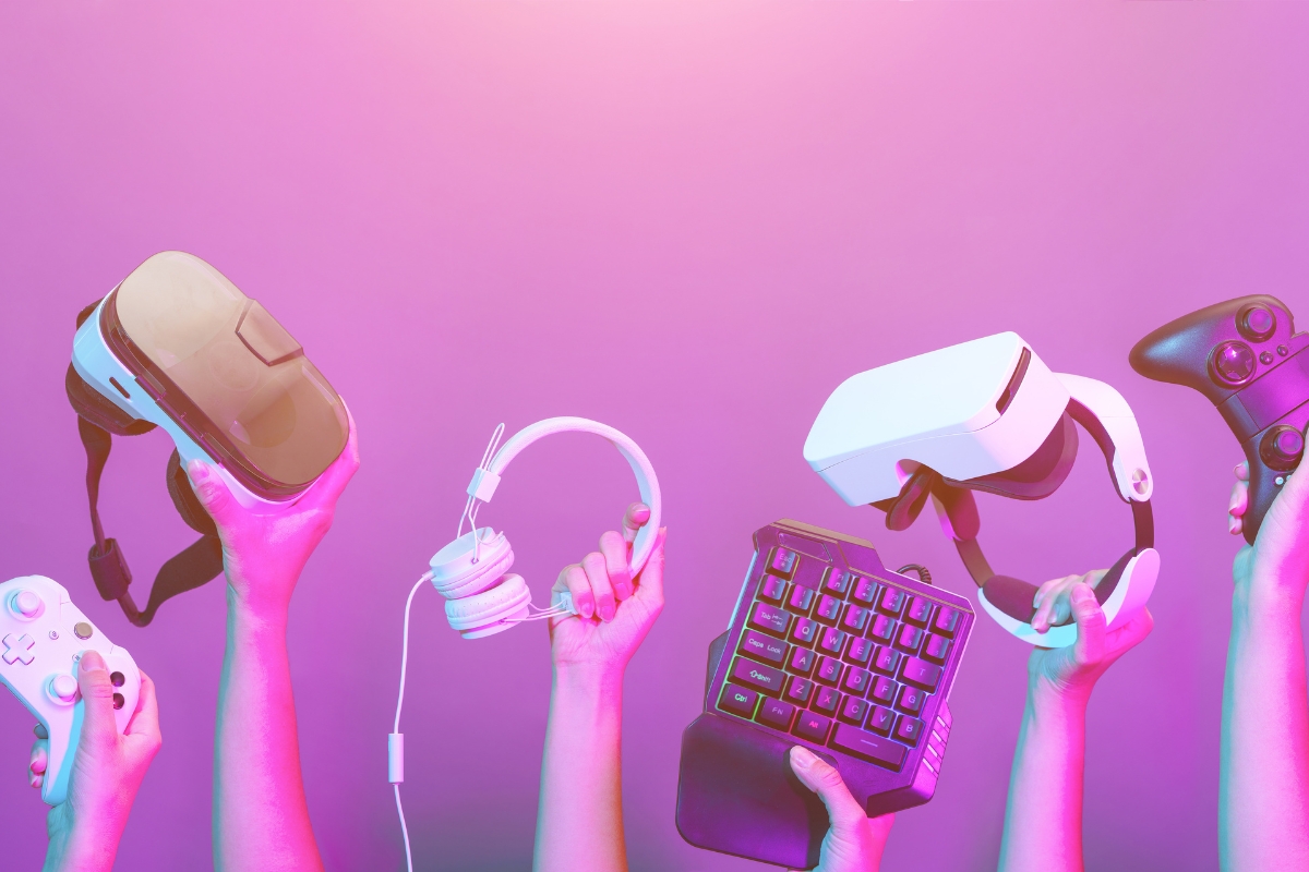 Gaming oprema za dečake – ruke drže VR naočare, slušalice, gejming tastaturu i kontrolere na roze-ljubičastoj pozadini.
