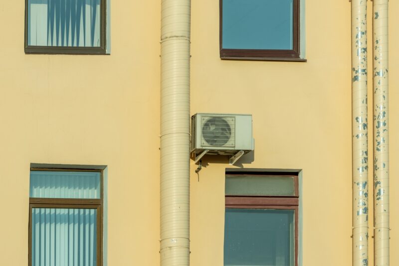 Spoljašnja jedinica klima uređaja postavljena na fasadi starije zgrade sa prozorima i cevima pored zida